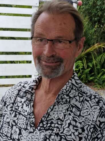 Un hombre mayor sonriente, con gafas y barba corta, está sentado al aire libre. Lleva una camisa estampada y está rodeado de vegetación y un fondo de madera blanca.