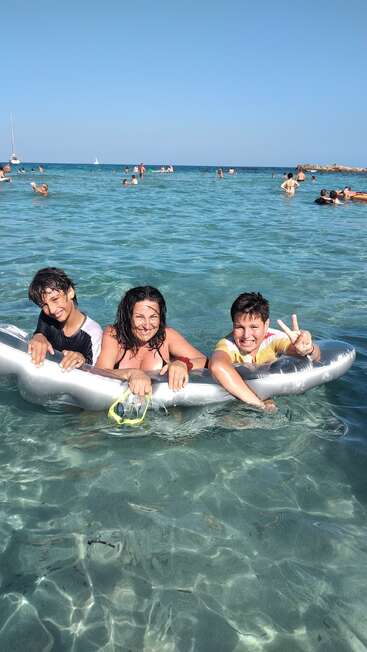 Três pessoas sorriem em uma boia em uma água azul clara, aproveitando um dia ensolarado na praia. Outras pessoas nadam nas proximidades, com barcos visíveis ao fundo. Diversão de verão.