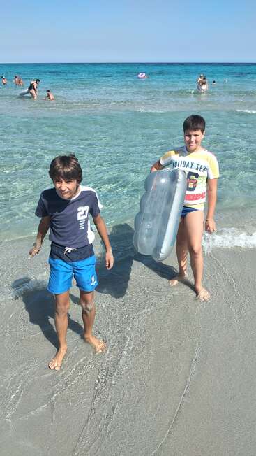 Dois meninos em pé em uma praia de areia perto de água azul clara, um deles segurando um colchão inflável. As pessoas nadam e brincam no mar sob um céu ensolarado.