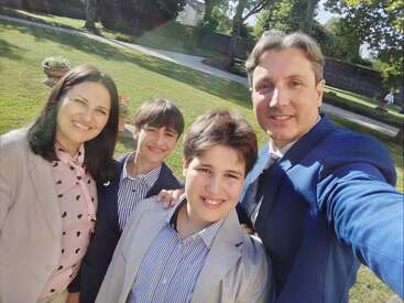 Una familia de cuatro miembros, vestida de gala, sonríe para hacerse un selfi al aire libre en un día soleado. Están muy juntos en un precioso jardín verde.