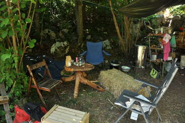 Esta imagem mostra um aconchegante acampamento ao ar livre com cadeiras, uma mesa de madeira, um fardo de feno e uma área de cozinha improvisada cercada por árvores e vegetação.