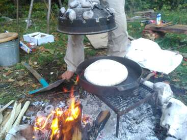 Jemand backt draußen Brot in einer gusseisernen Pfanne über dem Lagerfeuer. Er hebt den Deckel eines Dutch Ovens an, der mit heißen Kohlen bedeckt ist.