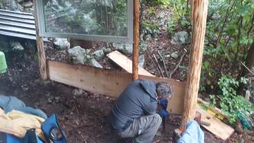 Eine Person in Outdoor-Kleidung baut in einem Waldgebiet einen Holzunterstand und befestigt Holz an senkrechten Pfosten neben einem großen Fenster.