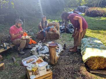 Drei Menschen genießen ein Beisammensein im Freien um eine Feuerstelle aus Stein herum, sitzen auf Strohballen und tauschen Essen und Gespräche aus – in einer rustikalen, handgefertigten Umgebung und bei strahlendem Sonnenschein.