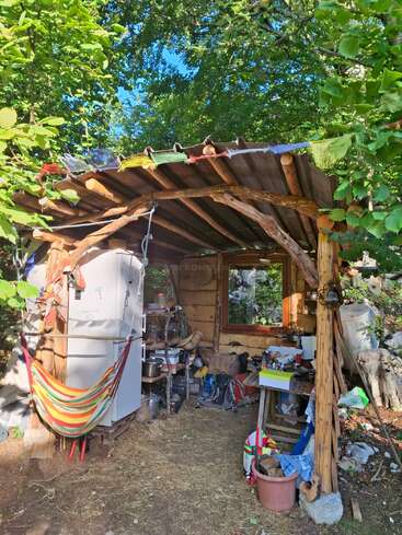 Inmitten üppiger Vegetation steht eine rustikale Holzhütte. Darin befinden sich ein Kühlschrank, eine Hängematte, Regale mit Töpfen und Vorräten, ein Tisch, ein Fenster und darüber bunte Gebetsfahnen.