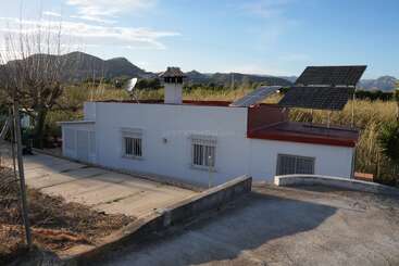 Uma casinha branca com janelas com grades, acabamentos vermelhos e painéis solares no telhado fica numa área rural cercada por montanhas e vegetação sob um céu azul.