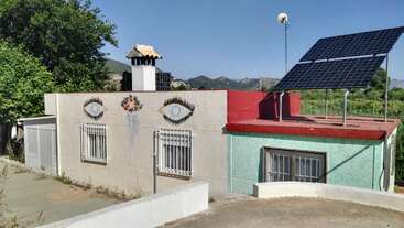 L'image représente un mur beige sur lequel sont peints deux yeux, accompagnés d'une mosaïque colorée et de deux fenêtres, le tout sur fond d'arbres et de montagnes.
