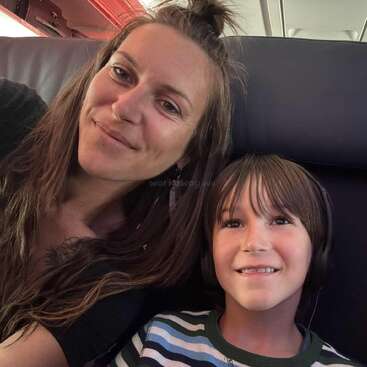 Uma mulher e um menino sorriem enquanto estão sentados juntos em um avião. O menino está usando fones de ouvido e uma camisa listrada. Eles parecem felizes.