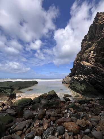 Una costa rocosa bajo un cielo azul intenso, con nubes dispersas que proyectan sombras. Acantilados escarpados y rocas cubiertas de musgo enmarcan una playa apartada y tranquila con suaves olas del mar. Una escena apacible.