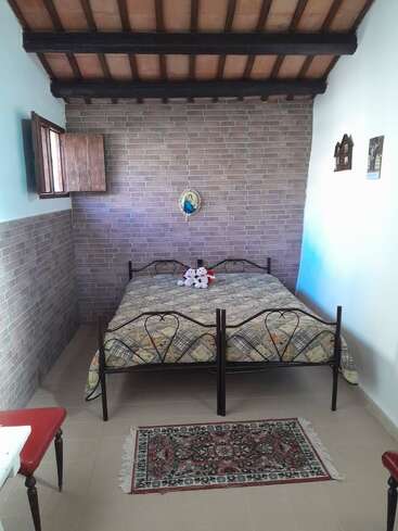 This image shows a cozy bedroom with a double bed, patterned quilt, brick walls, wooden ceiling, small rug, open window, and plush toys on the bed.