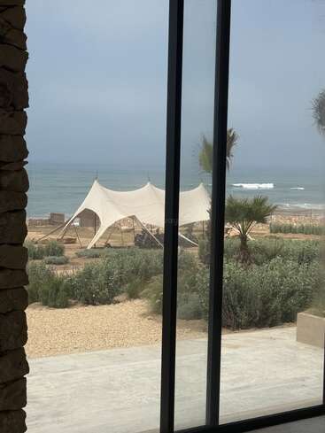 Vista a través de una puerta de cristal, una escena junto a la playa muestra una gran tienda blanca sobre suelo arenoso, exuberante vegetación, palmeras y suaves olas bajo un cielo nublado.