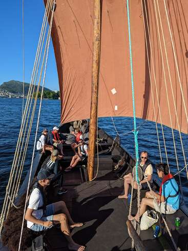 The image depicts a group of people on a sailboat, with a large brown sail and wooden mast, set against a serene body of water and a clear blue sky.