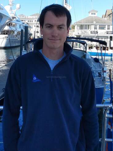 The image depicts a man standing on a dock, with boats in the background, wearing a navy blue quarter-zip sweatshirt with a logo on the left side of his chest.