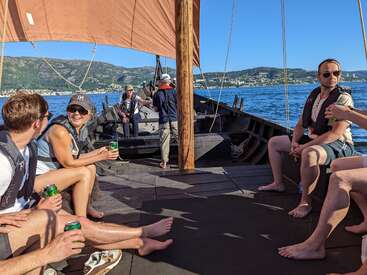 The image depicts a group of people on a boat in the water, enjoying drinks and each other\'s company on a sunny day with mountains in the background.