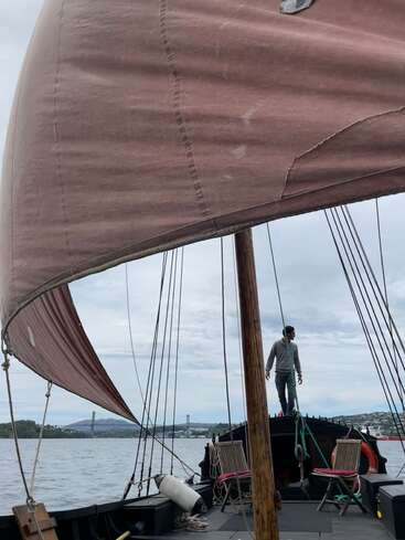 The image depicts a man standing on a boat with a large brown sail, set against a cloudy sky and a body of water in the background.