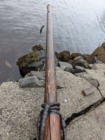 The image depicts a wooden pole with a rope wrapped around its base, situated on a rocky shoreline near the water\'s edge.