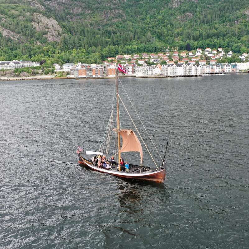 A small sailboat with a brown sail and wooden hull floats on a large body of water, with a town and mountains in the background.