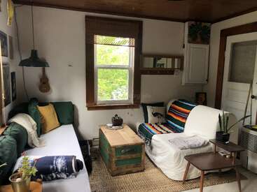 This cozy living room features a white couch with a colorful blanket, a wooden chest coffee table, green accents, window, plants, and bright natural light.