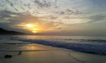 La imagen representa una serena escena playera al atardecer, con una suave ola que llega a la orilla y un cielo nublado.