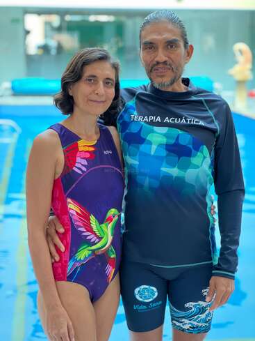 Una pareja sonriente posa junto a una piscina cubierta. Ella lleva un colorido bañador con motivos de colibríes y él, un traje azul de terapia acuática.