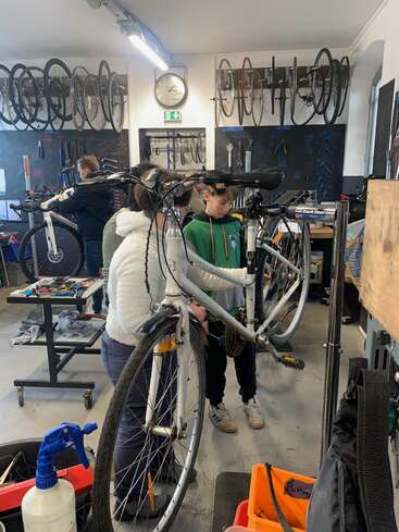 In a bicycle workshop, people, including children and adults, work together repairing bikes. Numerous wheels and tools hang on the walls, creating an organized workspace.