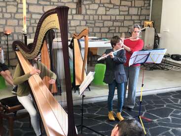 Three musicians perform indoors; two play the flute while one plays the harp. They stand near music stands, with stone walls and a candle behind them.
