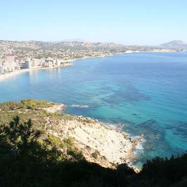 L'image représente une ville côtière avec une plage et des bâtiments, sur fond de montagnes et de ciel bleu clair.