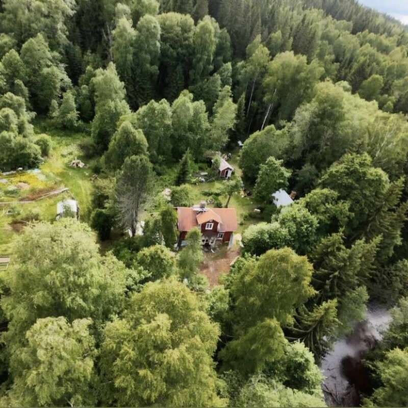 Cette image aérienne montre une maison rouge pittoresque nichée au milieu d'arbres denses et verdoyants, entourée d'une forêt, de jardins et d'un ruisseau, évoquant un havre de paix et la nature.
