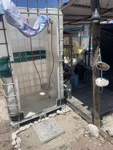 An outdoor makeshift shower made from a repurposed water tank, next to a rustic wooden shelter. Supplies and utility items are visible under the corrugated metal roof.