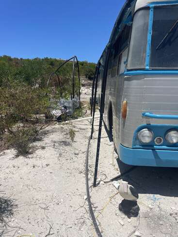 An old blue and silver bus is parked on sandy ground. Surrounding brush, pipes, water tanks, and a small bird statue add to the rustic, abandoned setting.