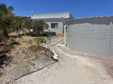 A large metal water tank stands beside a gray shed, surrounded by dry grass, sandy ground, scattered pipes, bushes, and a clear blue sky above.