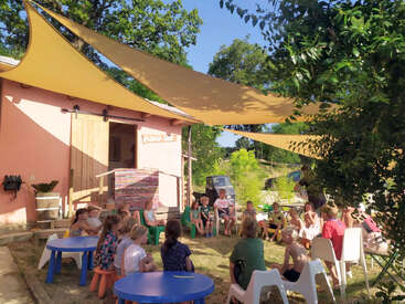 Eine Gruppe von Kindern sitzt draußen unter Sonnenschirmen, versammelt sich um bunte Tische und Stühle und nimmt an einer Aktivität in der Nähe eines rosa Gebäudes an einem sonnigen Tag teil.