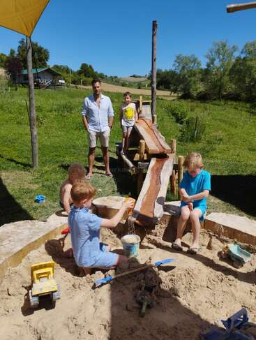 Kinder spielen an einem sonnigen Tag im Freien auf einem Sandplatz mit Spielzeug und Wasser, im Hintergrund sind grüne Wiesen und Bäume zu sehen, während zwei Erwachsene auf sie aufpassen.