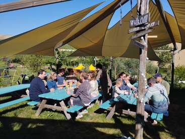 Die Leute sitzen an Picknicktischen unter gelben Sonnensegeln und genießen Getränke, gute Gespräche und die Sonne. Die Atmosphäre ist entspannt, man ist draußen, umgeben von Bäumen und Gras, und es herrscht eine ausgelassene Stimmung.