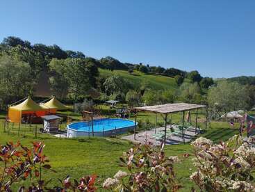 Eine lebhafte Landschaftsszene zeigt einen runden Swimmingpool, gelbe Zelte, Liegestühle im Schatten, grüne Hügel, üppige Bäume, blauen Himmel und blühende Pflanzen im Vordergrund.