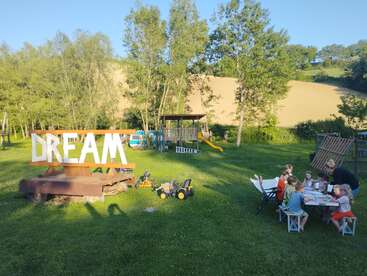 Eine Gruppe von Kindern und ein Erwachsener sitzen draußen an einem Tisch und basteln, in der Nähe eines großen „DREAM“-Schilds, umgeben von grünem Gras, Bäumen und Spielgeräten.