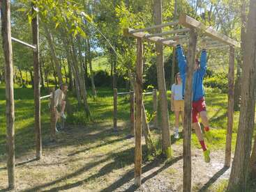 Drei Leute vergnügen sich auf einem Outdoor-Hindernisparcours in einem sonnigen, grünen Park. Eine Person schaukelt am Klettergerüst; zwei andere schauen zu und warten in der Nähe zwischen hohen Bäumen.