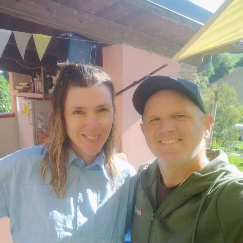 Ein lächelndes Paar macht an einem sonnigen Tag draußen ein Selfie, mit grüner Landschaft und bunten Wimpeln im Hintergrund, was eine fröhliche, entspannte Atmosphäre schafft.