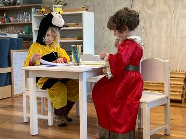 Duas crianças fantasiadas — uma vestida de abelha e a outra com um vestido vermelho de princesa — estão sentadas à mesa, desenhando com canetas coloridas em um quarto aconchegante.