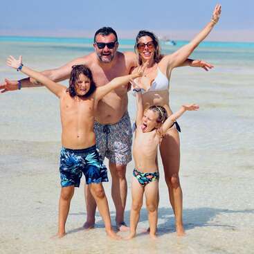 Eine glückliche vierköpfige Familie steht im flachen Strandwasser, lächelt mit weit geöffneten Armen und genießt einen sonnigen Tag am Meer, mit Badeanzug und Sonnenbrille.