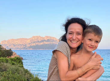 The image depicts a woman and a young boy embracing on a cliff overlooking the ocean, with a mountain range visible in the background under a clear blue sky.