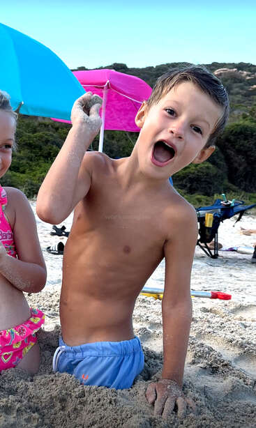 The image depicts a young boy with his mouth open, sitting in the sand on a beach, with a pink and blue umbrella behind him and a girl to his left.