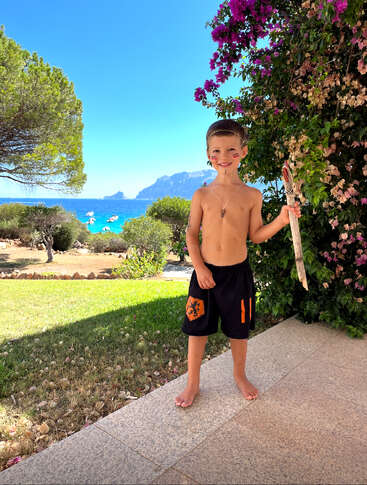 The image depicts a young boy standing on a stone patio, holding a stick, with a serene ocean view and lush greenery in the background on a sunny day.