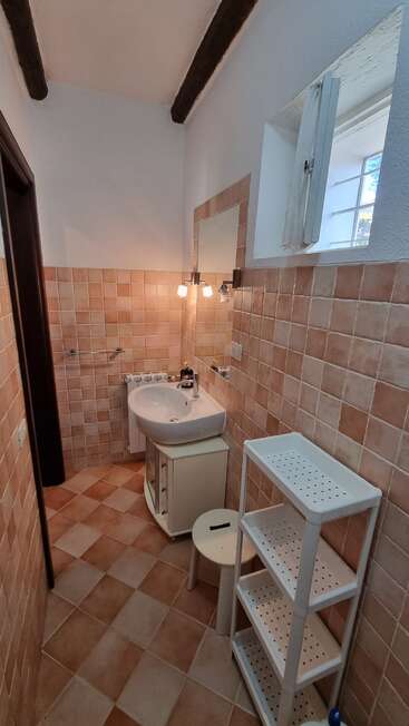 The image depicts a compact bathroom with a sink, tiled walls, and a window, featuring a small white shelving unit and a stool, with a mirror above the sink.
