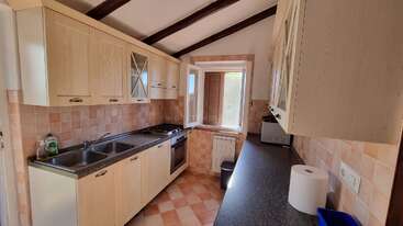 The image depicts a kitchen with a sink, stove, and refrigerator, featuring a wooden table and chairs, with a window and a door visible in the background. The room is well-lit and organized.