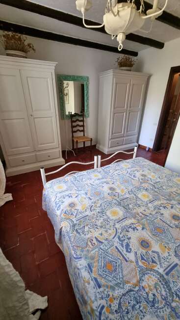 The image depicts a bedroom with white walls, red tile floors, and white furniture, featuring a bed with a blue and yellow patterned cover and a mirror on the wall.
