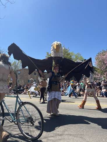 Bei einer lebhaften Parade trägt jemand ein riesiges Adlerkostüm mit ausgebreiteten Flügeln. Die Zuschauer schauen zu, während andere in kreativen Outfits und auf einem Fahrrad in der Nähe auftauchen.
