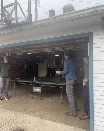 Drei Männer sind in einer Garage mit einem Tischtennistisch; einer spielt, während die anderen zuschauen. Über dem offenen Garagentor ist ein Basketballkorb angebracht.