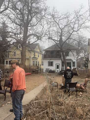Zwei Menschen stehen in einem Hinterhof mit Gartenstühlen, kahlen Bäumen und zwei Häusern im Hintergrund. Es sieht bewölkt aus, mit braunem Gras und einer entspannten Atmosphäre.