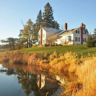 The image depicts a serene white house with a red chimney, situated on a hill overlooking a tranquil pond and surrounded by trees and golden grass.